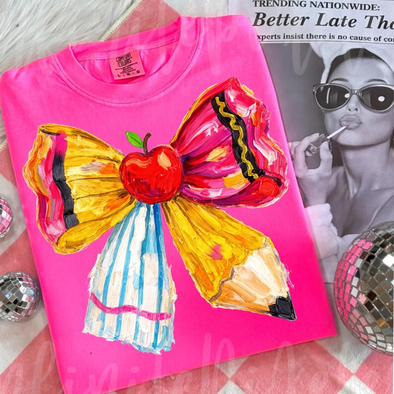 May include: A bright pink sweatshirt featuring a colorful, artistic design of a pencil-shaped bow with a red apple at the center. The bow has yellow, pink, and black accents. The sweatshirt is folded on a red and white checkered surface.