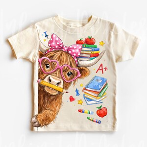 Pu&ograve; includere: T-shirt color crema con un cartone animato di una mucca delle Highlands con occhiali a forma di cuore e un fiocco a pois. La mucca tiene una matita ed &egrave; circondata da libri, mele e pastelli. La maglietta ha la scritta "A+".