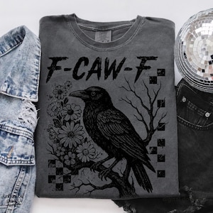 Puede incluir: Camiseta gris oscuro con un dise&ntilde;o negro de un cuervo posado en una rama, rodeado de flores y el texto "F-CAW-F". El dise&ntilde;o incluye un patr&oacute;n de cuadros y ramas de &aacute;rboles desnudas.