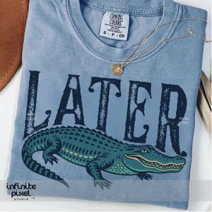 Pode incluir: Uma camiseta azul claro com a palavra "LATER" impressa em uma fonte de estilo vintage. Um jacar&eacute; verde com um sorriso de dentes est&aacute; impresso abaixo da palavra.
