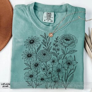 May include: A seafoam green t-shirt with a floral design. The design features various flowers and leaves in black ink. The shirt has the text "ORIGINAL DESIGNER" and "infinite pixel studio". A gold necklace with a pendant rests on the shirt.