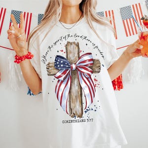 K&ouml;nnte beinhalten: Wei&szlig;es T-Shirt mit einem Holzkreuz und einer rot-wei&szlig;-blauen Schleife. Das Kreuz ist mit Sternen und dem Text "Where the spirit of the Lord there is freedom" und "Corinthians 3:17" verziert.