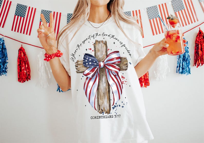 K&ouml;nnte beinhalten: Wei&szlig;es T-Shirt mit einem Holzkreuz und einer rot-wei&szlig;-blauen Schleife. Das Kreuz ist mit Sternen und dem Text "Where the spirit of the Lord there is freedom" und "Corinthians 3:17" verziert.