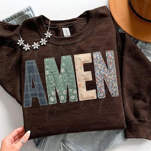 May include: A brown sweatshirt with the word "AMEN" appliqued across the front. The letters are made of various patterned fabrics, including plaid, floral, and gingham. A silver necklace with flower-shaped pendants rests on the sweatshirt.