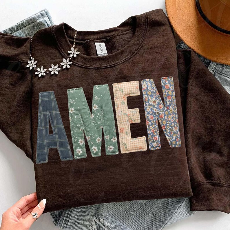 May include: A brown sweatshirt with the word "AMEN" appliqued across the front. The letters are made of various patterned fabrics, including plaid, floral, and gingham. A silver necklace with flower-shaped pendants rests on the sweatshirt.