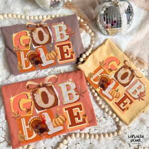 May include: Three fall-themed t-shirts in brown, peach, and yellow, each with the word "GOBBLE" in a colorful, layered design. The design includes a turkey, pumpkins, and fall leaves. A decorative bow is at the top of each shirt.