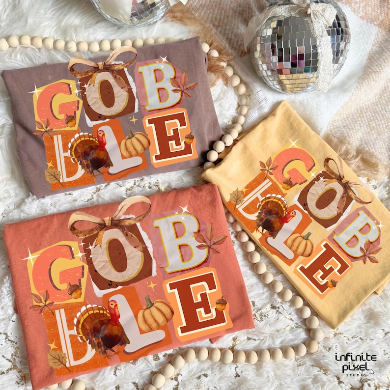 May include: Three fall-themed t-shirts in brown, peach, and yellow, each with the word "GOBBLE" in a colorful, layered design. The design includes a turkey, pumpkins, and fall leaves. A decorative bow is at the top of each shirt.
