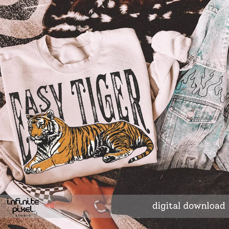 May include: Cream-coloured sweatshirt with the text "EASY TIGER" in black, featuring a tiger illustration in orange and black. A light blue denim jacket with flame designs is visible. The image also includes a checkered shoe.