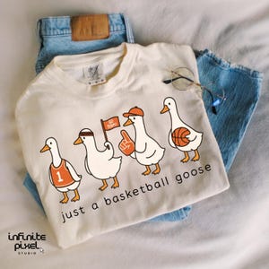 May include: Cream-coloured t-shirt featuring the text "just a basketball goose" and a graphic of four cartoon geese in basketball attire. The shirt is paired with blue jeans and glasses.