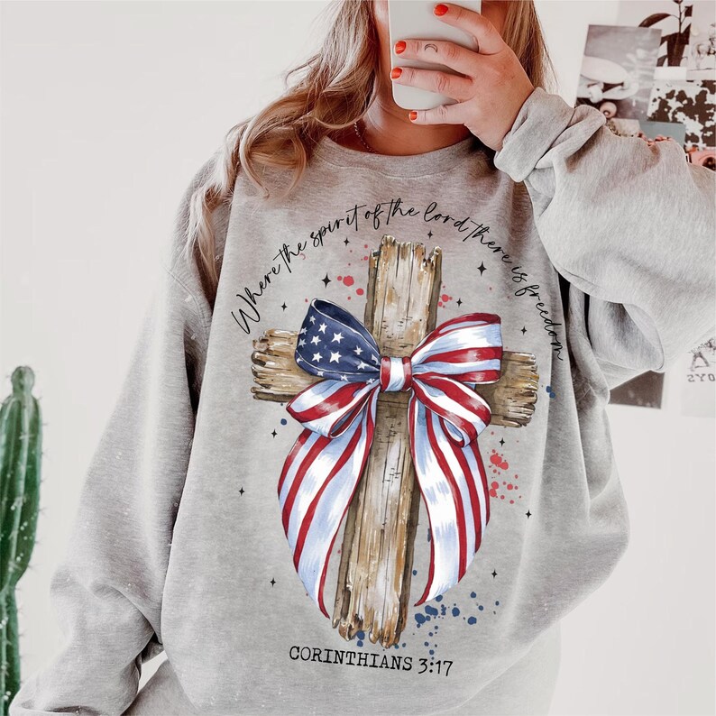 K&ouml;nnte beinhalten: Grauer Pullover mit einem Holzkreuz und einer rot-wei&szlig;-blauen Schleife. Das Kreuz ist mit Sternen und dem Text "Where the spirit of the Lord there is freedom" und "Corinthians 3:17" verziert.