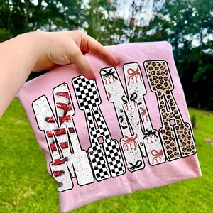 May include: A pale pink t-shirt featuring the word "MAMA" in a large, bold font. Each letter is decorated differently: red glitter, a black and white checkerboard, baseballs with bows, and leopard print. The shirt is held up by a hand.
