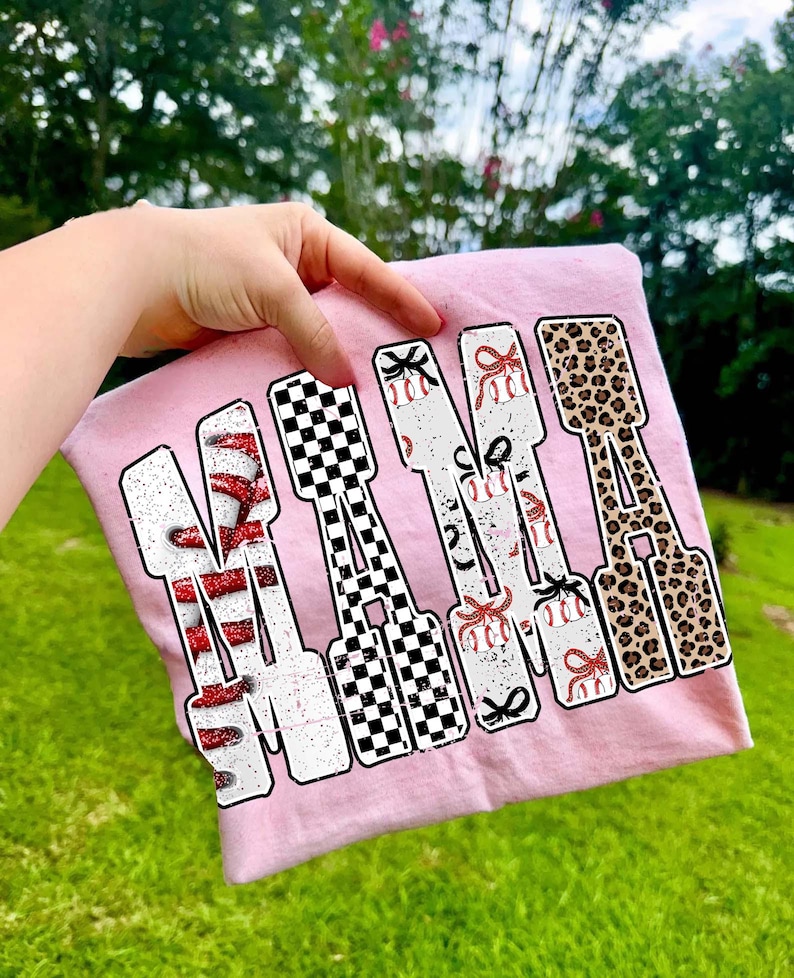 May include: A pale pink t-shirt featuring the word "MAMA" in a large, bold font. Each letter is decorated differently: red glitter, a black and white checkerboard, baseballs with bows, and leopard print. The shirt is held up by a hand.