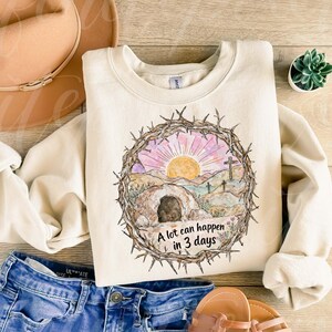 May include: Cream-colored sweatshirt with a watercolor graphic of a sunrise over a landscape with a tomb and crosses, encircled by a crown of thorns. The text "A lot can happen in 3 days" is below the graphic. The sweatshirt is paired with blue jeans and sandals.