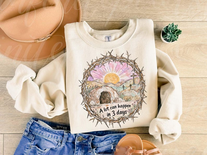 May include: Cream-colored sweatshirt with a watercolor graphic of a sunrise over a landscape with a tomb and crosses, encircled by a crown of thorns. The text "A lot can happen in 3 days" is below the graphic. The sweatshirt is paired with blue jeans and sandals.