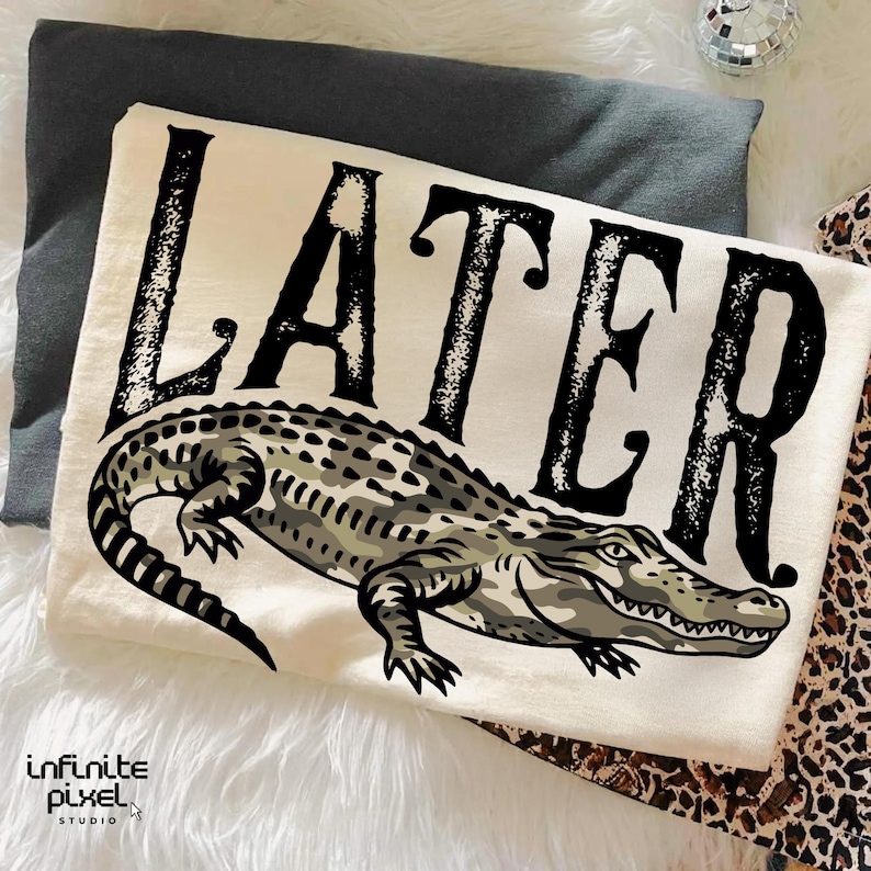 Peut inclure: T-shirt cr&egrave;me avec une illustration d&eacute;taill&eacute;e d'un alligator sous le mot "LATER" en lettres noires en gras. L'alligator est rendu dans des tons de gris et de vert. Le t-shirt est pli&eacute; sur une surface blanche.