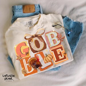 May include: Cream-colored t-shirt with the word "GOBBLE" in colorful, block letters, featuring a turkey, pumpkin, and fall leaves. The shirt is laid on top of blue jeans, with a pair of glasses resting on the shirt.