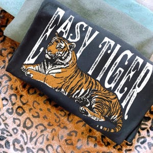 May include: A black t-shirt with the words "EASY TIGER" in white, featuring a detailed illustration of a tiger in shades of brown and orange. The shirt is folded, with other fabric visible in the background.