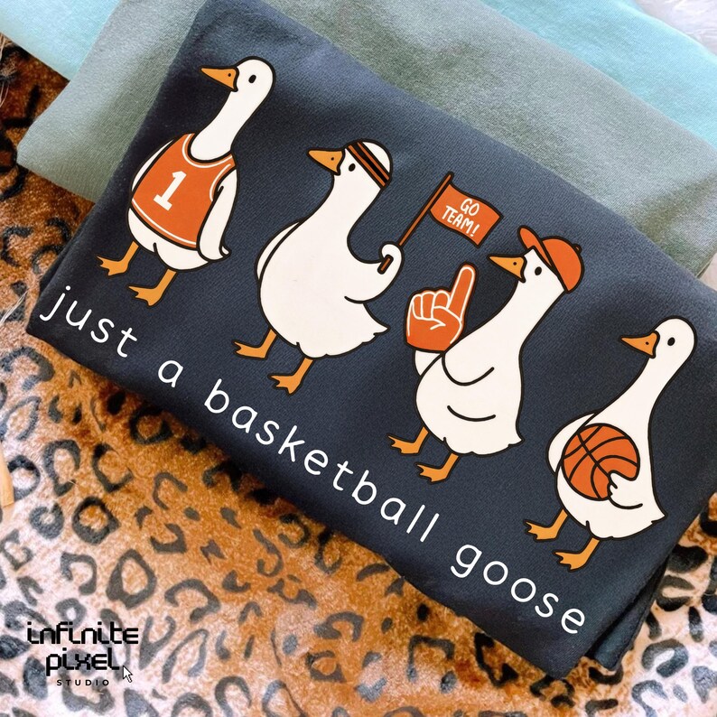 May include: Dark grey t-shirt with a basketball theme, featuring four cartoon geese. The geese are wearing a jersey, headband, holding a flag, and a basketball. The text "just a basketball goose" is printed on the shirt.