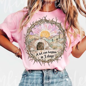 May include: Pink t-shirt featuring a watercolor design of a sunrise over a landscape with a tomb and crosses, encircled by a crown of thorns. The text "A lot can happen in 3 days" is below the image.