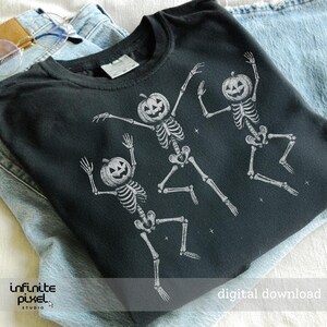 May include: Black crew neck t-shirt featuring three dancing skeletons with pumpkin heads. The skeletons are white and the shirt has the text "digital download". The shirt is folded on top of a pair of blue jeans.