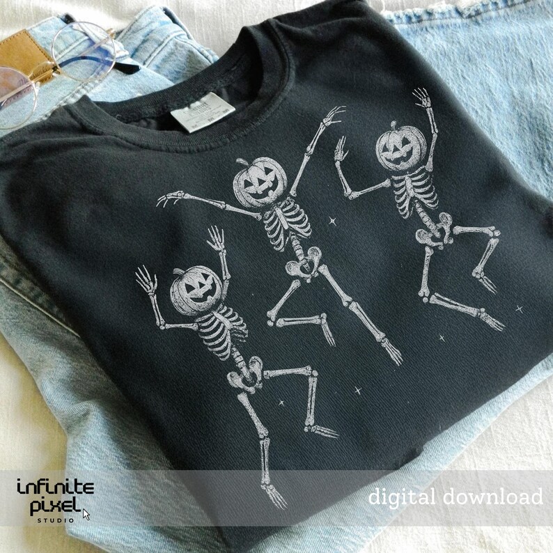 May include: Black crew neck t-shirt featuring three dancing skeletons with pumpkin heads. The skeletons are white and the shirt has the text "digital download". The shirt is folded on top of a pair of blue jeans.