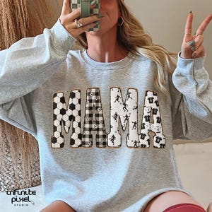 May include: A light gray sweatshirt with the word "MAMA" in large, decorative letters. The letters are filled with soccer ball patterns, a black and white gingham pattern, soccer player silhouettes, and soccer jersey designs. The sweatshirt has a crew neck.
