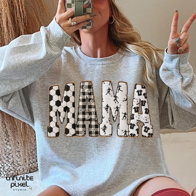 May include: A light gray sweatshirt with the word "MAMA" in large, decorative letters. The letters are filled with soccer ball patterns, a black and white gingham pattern, soccer player silhouettes, and soccer jersey designs. The sweatshirt has a crew neck.