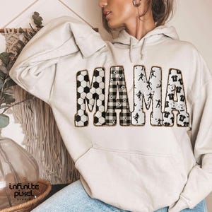 May include: A light beige hoodie with the word "MAMA" in large, bold letters across the front. The letters are decorated with soccer ball patterns, plaid, and soccer player silhouettes. The hoodie has a kangaroo pocket and a drawstring hood.
