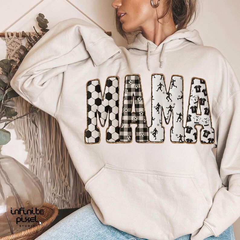 May include: A light beige hoodie with the word "MAMA" in large, bold letters across the front. The letters are decorated with soccer ball patterns, plaid, and soccer player silhouettes. The hoodie has a kangaroo pocket and a drawstring hood.
