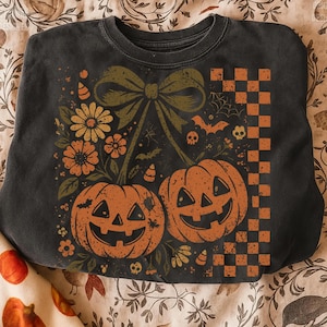 May include: Dark gray sweatshirt featuring a Halloween-themed design. The graphic includes two orange jack-o'-lanterns, a large green bow, flowers, bats, and a checkered pattern. The design has a vintage, distressed look.