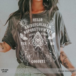 May include: Dark grey t-shirt featuring a white Ouija board design. The design includes the alphabet, numbers, sun, moon, butterflies, and the words "Hello", "Yes", "No", and "Goodbye".
