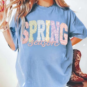 May include: A light blue t-shirt with the words "SPRING season" in colorful patchwork letters. The letters are in various patterns, including floral and gingham. The shirt is worn with denim shorts and cowboy boots.