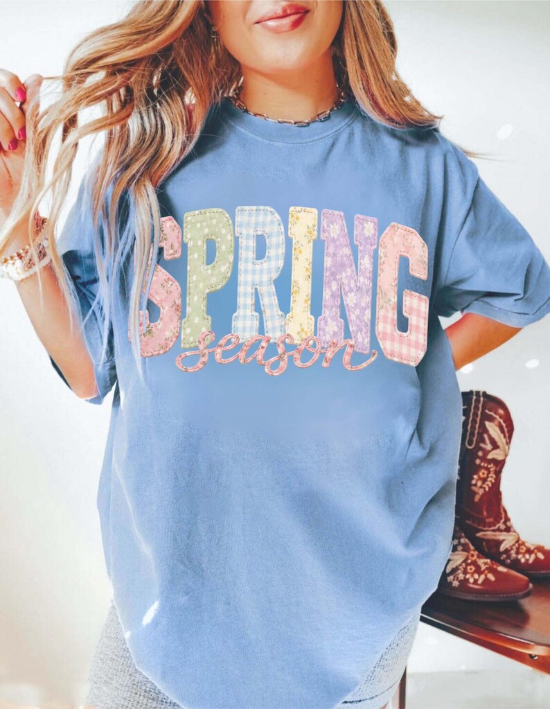 May include: A light blue t-shirt with the words "SPRING season" in colorful patchwork letters. The letters are in various patterns, including floral and gingham. The shirt is worn with denim shorts and cowboy boots.