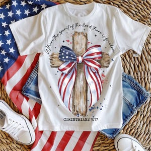 K&ouml;nnte beinhalten: Wei&szlig;es T-Shirt mit einer rot-wei&szlig;-blauen Schleife, die um ein Holzkreuz gebunden ist. Der Text "Where the spirit of the Lord there is freedom" ist auf dem Shirt aufgedruckt, sowie der Text "Corinthians 3:17".