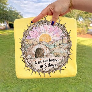 May include: Yellow tote bag featuring a watercolor illustration of a sunrise over a landscape with three crosses and a tomb. The image is framed by a crown of thorns. Text reads "A lot can happen in 3 days."