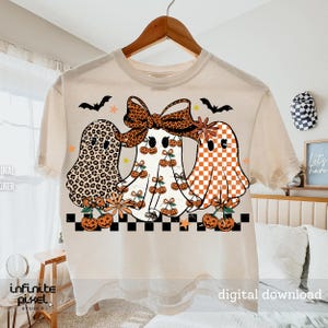 May include: Cream-colored t-shirt featuring three cartoon ghosts with unique patterns: leopard print, cherries, and orange checkered. The ghosts are adorned with bows and surrounded by bats, stars, and pumpkins. The shirt has a black and white checkered border.