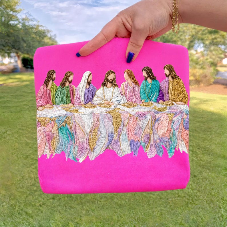 May include: A hot pink clutch featuring an embroidered depiction of The Last Supper. The embroidery showcases figures in colorful robes seated at a table with a detailed, textured tablecloth. The clutch is held up by a hand.