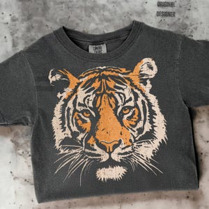 May include: A charcoal gray t-shirt featuring a distressed tiger face graphic in orange and cream. The shirt has a crew neck and short sleeves. The tag reads "Comfort Colors" and the text "ORIGINAL DESIGNER" is at the top.