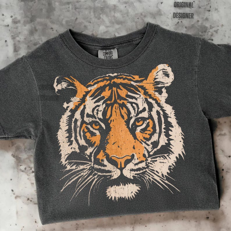 May include: A charcoal gray t-shirt featuring a distressed tiger face graphic in orange and cream. The shirt has a crew neck and short sleeves. The tag reads "Comfort Colors" and the text "ORIGINAL DESIGNER" is at the top.