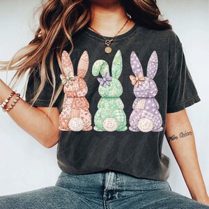 May include: Dark gray t-shirt featuring three patchwork bunny rabbits in pastel colors. Each bunny has a bow and a fluffy tail. The bunnies are in shades of pink, green, and purple, with floral and gingham patterns.