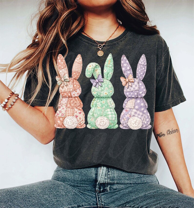 May include: Dark gray t-shirt featuring three patchwork bunny rabbits in pastel colors. Each bunny has a bow and a fluffy tail. The bunnies are in shades of pink, green, and purple, with floral and gingham patterns.