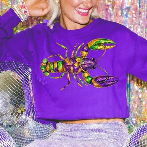 May include: A purple sweatshirt featuring a colorful illustration of a lobster. The lobster design incorporates shades of green, gold, and purple. The sweatshirt is a solid purple color.