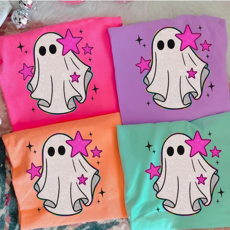 May include: Four colorful t-shirts: pink, purple, orange, and teal. Each shirt features a white ghost graphic with black eyes and a pink star. Additional pink stars and black sparkles surround the ghost design. A fun and festive design for Halloween.