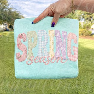 May include: A light blue square-shaped decorative item with the words "SPRING season" in colorful, patterned letters. The letters are in pink floral, green polka dot, blue gingham, yellow floral, purple floral, and pink gingham patterns.