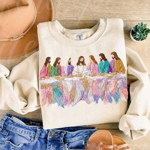 May include: Cream-colored sweatshirt featuring an embroidered depiction of the Last Supper. The design showcases figures in colorful robes seated at a table, with a detailed, multi-colored lower border. The sweatshirt is paired with blue jeans and sandals.