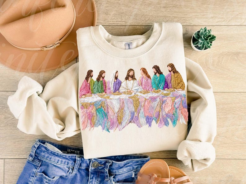 May include: Cream-colored sweatshirt featuring an embroidered depiction of the Last Supper. The design showcases figures in colorful robes seated at a table, with a detailed, multi-colored lower border. The sweatshirt is paired with blue jeans and sandals.