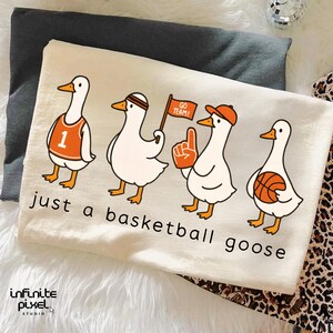 May include: Cream-coloured t-shirt with a cartoon design of four geese in basketball attire. The geese are wearing a jersey, headband, hat, and holding a flag, foam finger, and basketball. The text "just a basketball goose" is printed below.