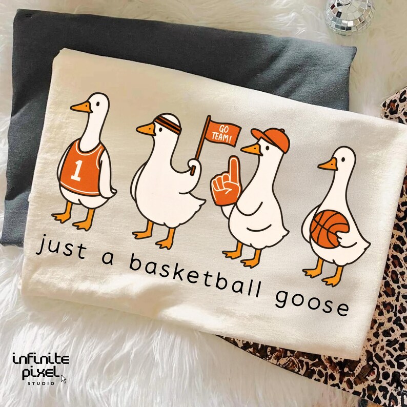 May include: Cream-coloured t-shirt with a cartoon design of four geese in basketball attire. The geese are wearing a jersey, headband, hat, and holding a flag, foam finger, and basketball. The text "just a basketball goose" is printed below.