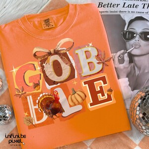 May include: Orange Comfort Colors t-shirt with a Thanksgiving-themed graphic. The word "GOBBLE" is spelled out in colorful letters with a turkey, pumpkin, and fall leaves. The shirt also has the text "infinite pixel studio".
