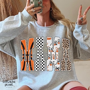 Puede incluir: Una sudadera gris claro con la palabra "MAMA" en letras grandes y decorativas. La "M" es naranja con un dise&ntilde;o de baloncesto, la primera "A" es a cuadros blancos y negros, la segunda "M" tiene un dise&ntilde;o de calabaza y lazo, y la &uacute;ltima "A" es de estampado de leopardo.